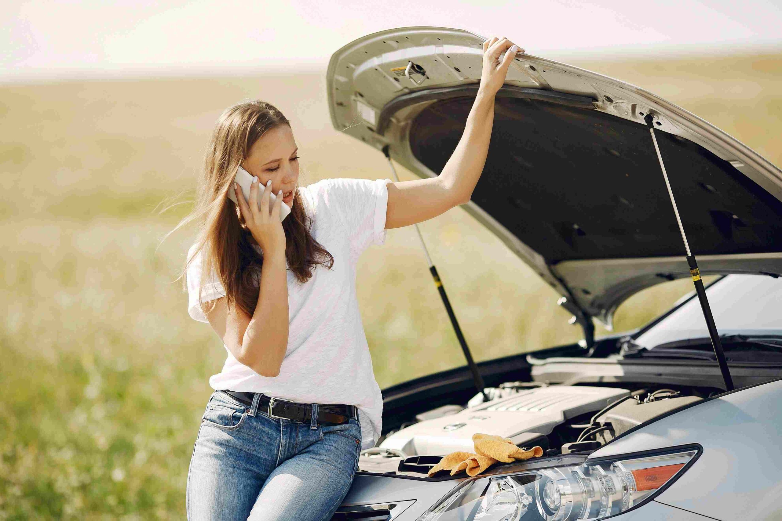 A woman uses a cell phone while leaning on the open hood of a car.