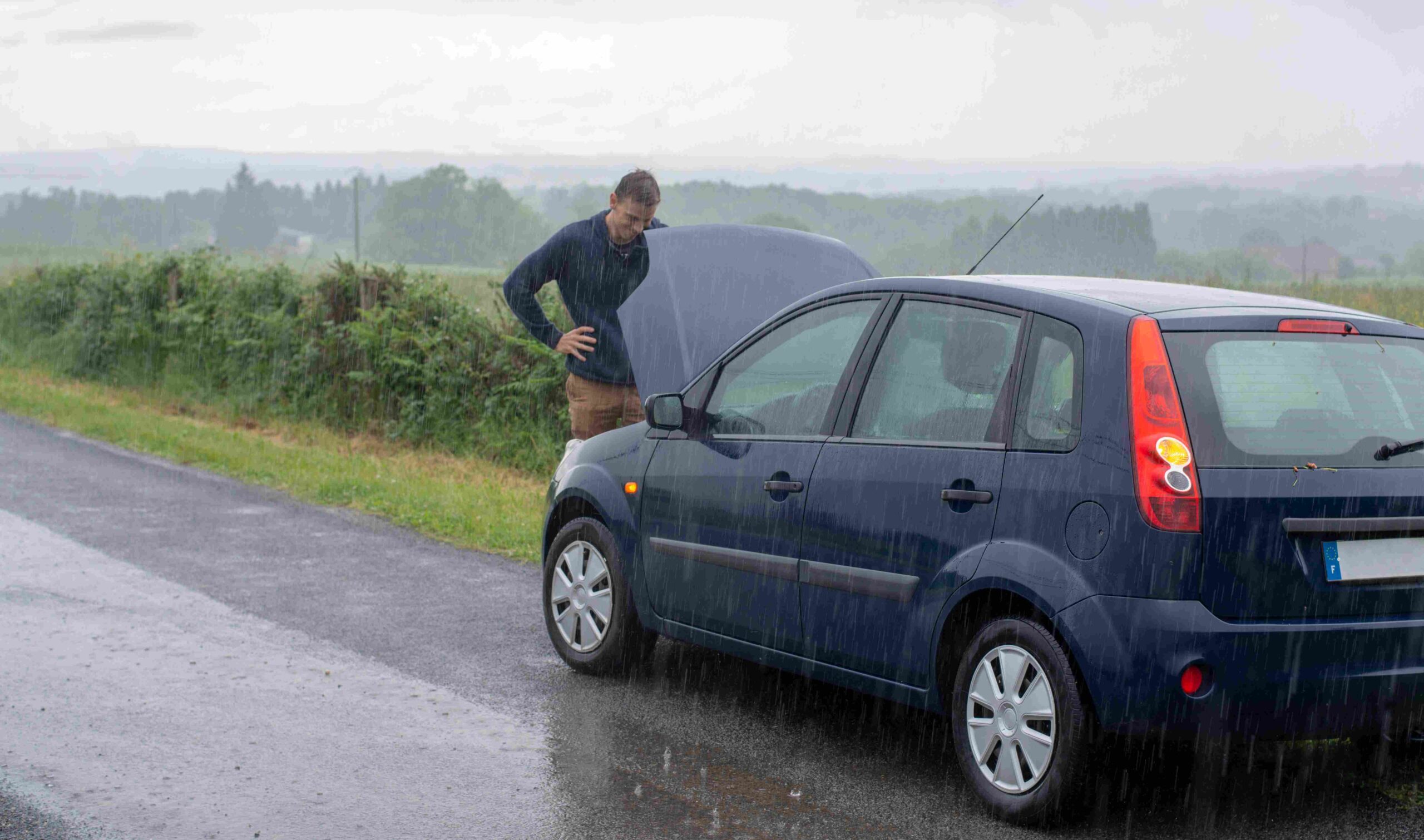 A man stands in front of a car broken down in the rain.